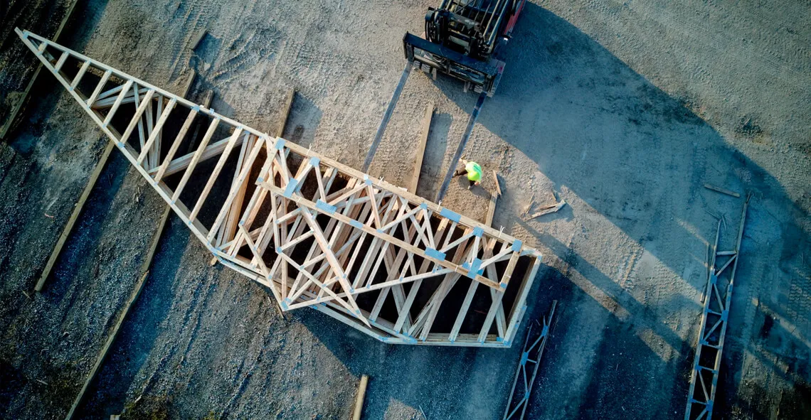Overhead view of a worker beside a large wood roof truss on a gravel yard